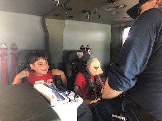 Two kids sitting inside a vehicle, with an adult in uniform talking to them.