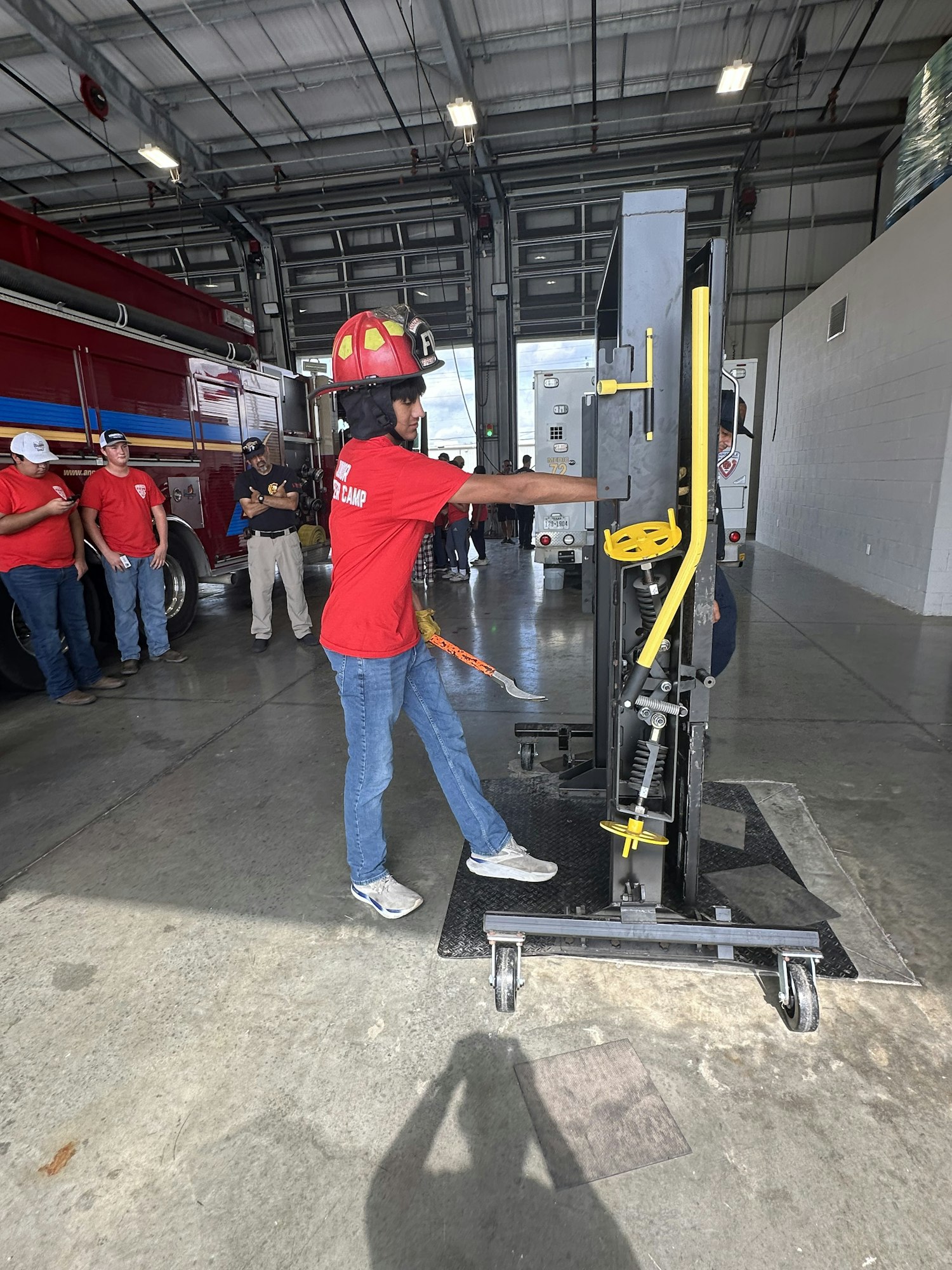 A person in a red shirt and helmet is using a firefighting training machine indoors, with others observing.