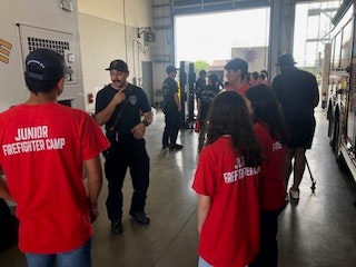 A firefighter talks to kids wearing "Junior Firefighter Camp" shirts in a garage or station.