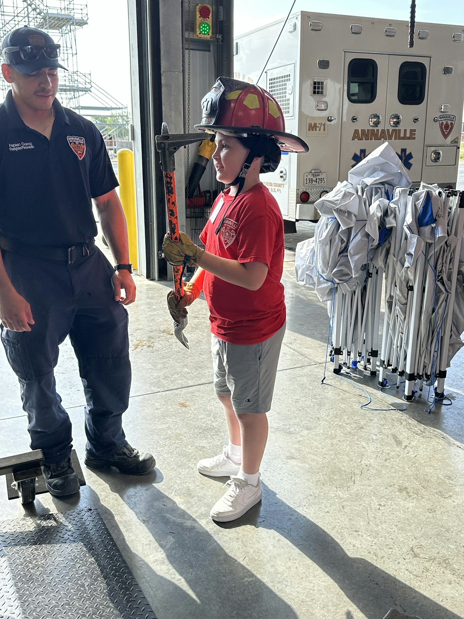 A child wearing a firefighter helmet holds a tool, standing next to an adult. An ambulance and equipment are in the background.