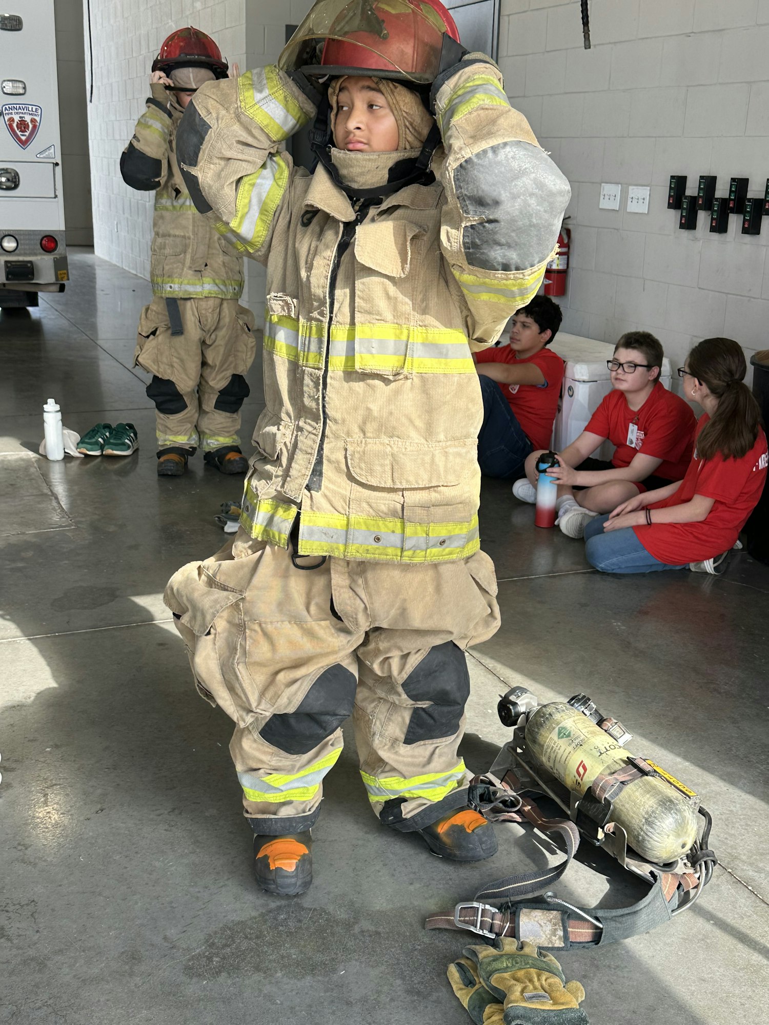 People in firefighting gear, with helmets and equipment, inside a station. Others in red shirts are sitting nearby.