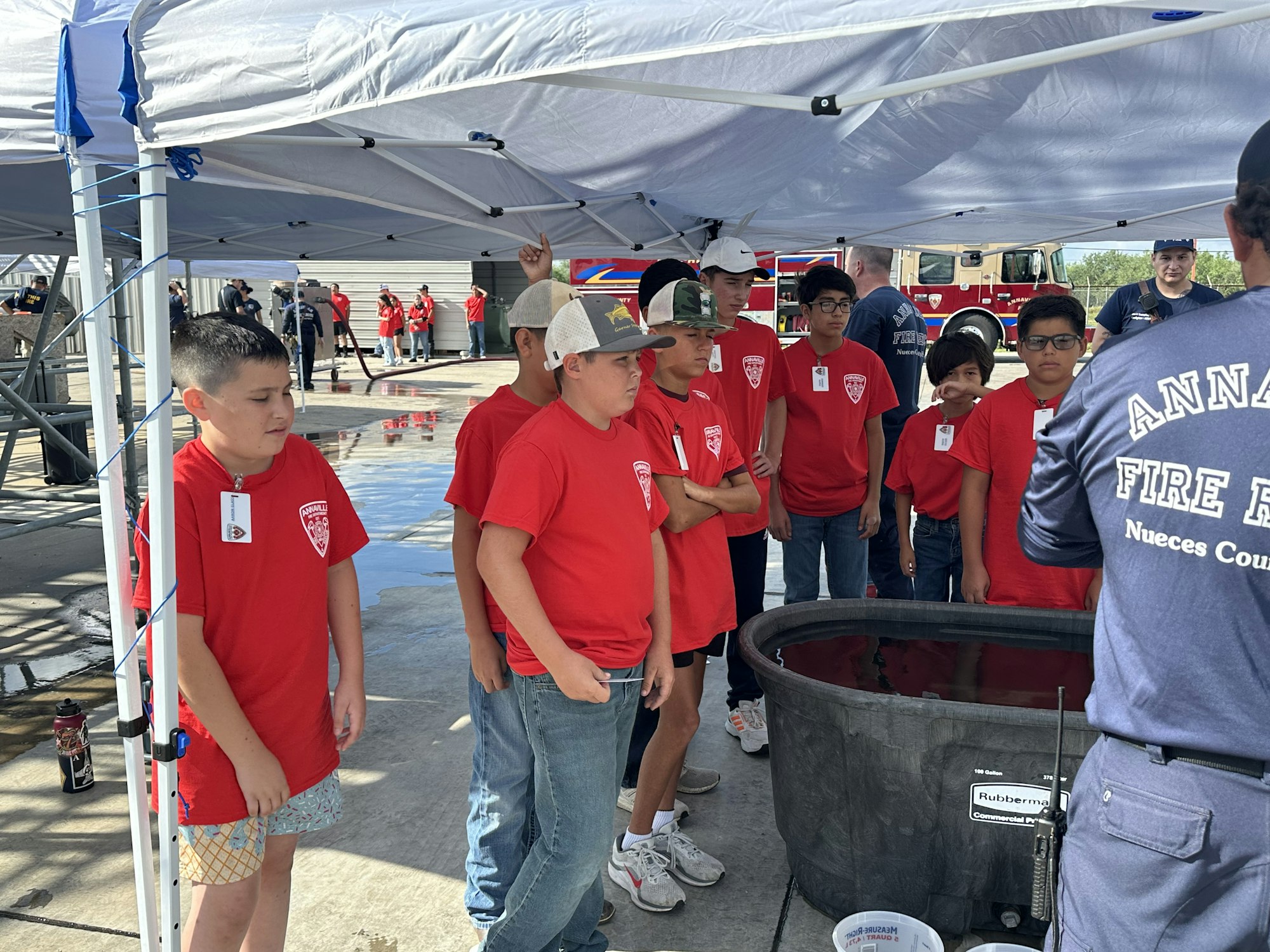 A group of children in red shirts observing a demonstration by fire department personnel under a canopy.