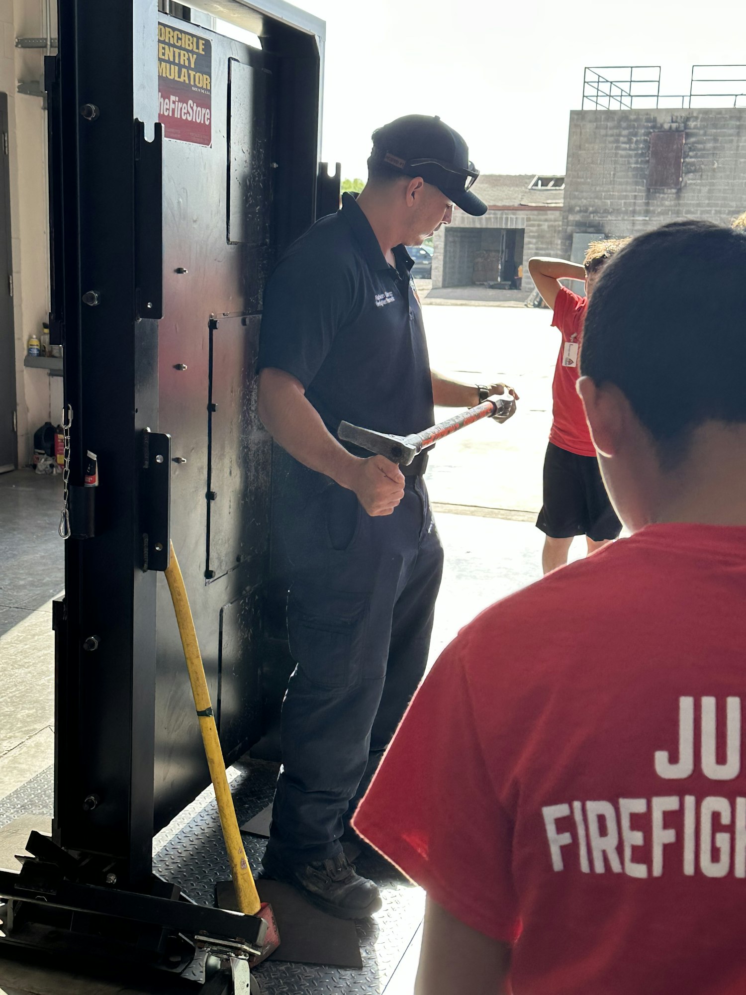 Person demonstrating with a tool near a forcible entry simulator. Others present, labeled "Junior Firefighter."