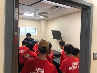 A group wearing "Junior Firefighter" shirts in a small room, facing a person at the front, with a wall-mounted TV.