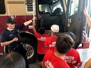 A firefighter shows kids in "Junior Firefighter Camp" shirts the inside of a fire truck.