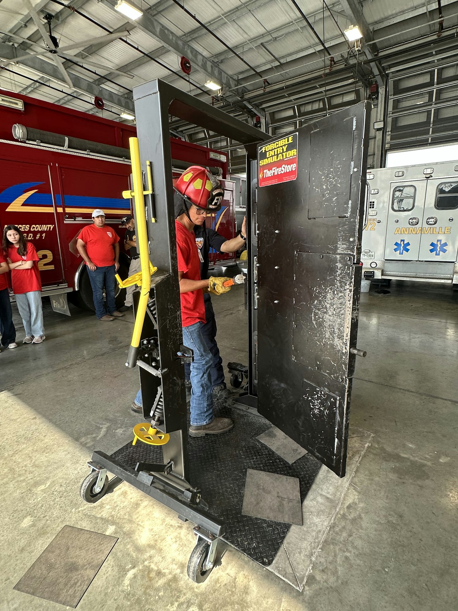 Firefighter in a red helmet using a tool on a black metal door frame, in a garage with an ambulance in the background.
