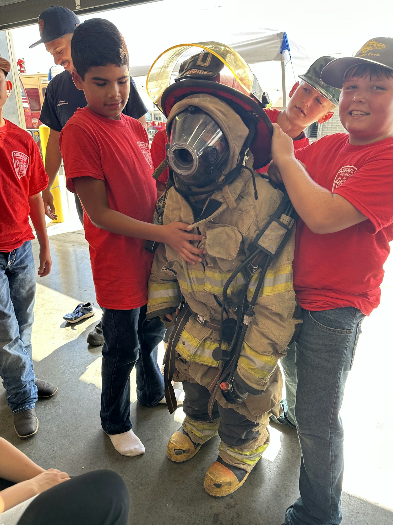Kids in red shirts helping a child try on firefighting gear, including a helmet and suit.