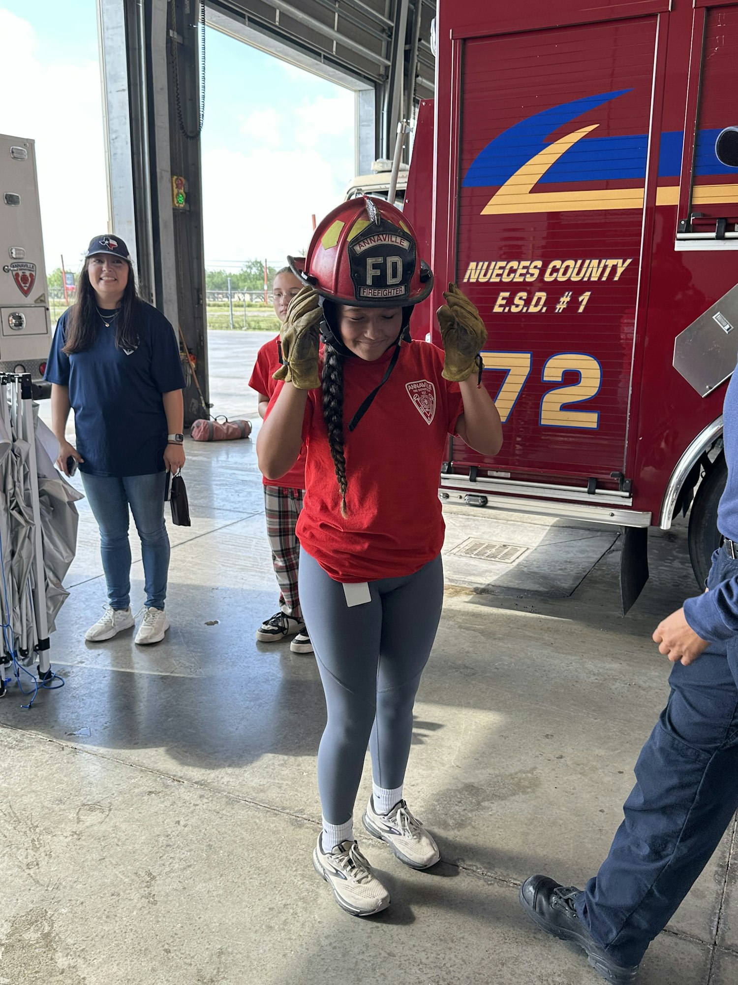 A person wearing a firefighter helmet in front of a fire truck, with others nearby in a garage setting.