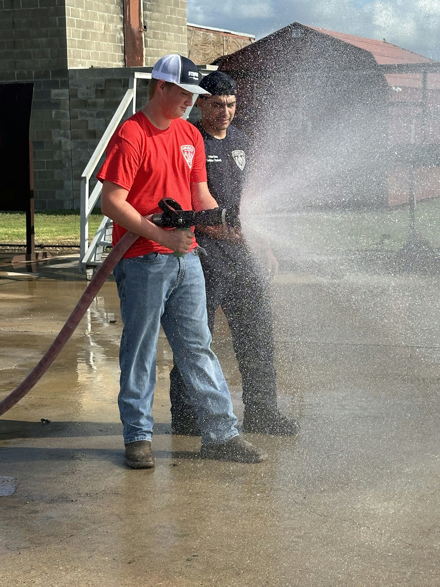 Two people using a fire hose spraying water on a sunny day, one wearing red and white, the other in black.