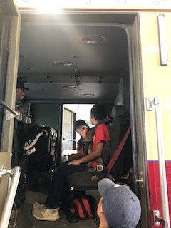 Inside view of a vehicle with two people sitting in seats wearing seat belts, one wearing a hat.
