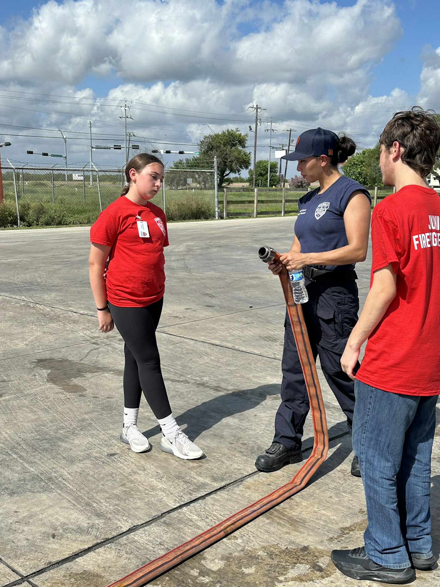 Three people outdoors, two in red shirts, one with a hose, discussing something on a sunny day.