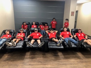 A group of kids wearing red shirts and caps sitting and relaxing on black recliners in a room.