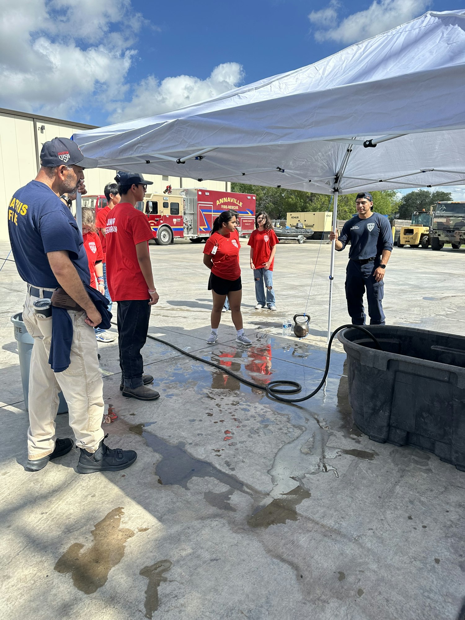People standing outdoors under a canopy, near a fire truck and equipment, possibly at a fire department or training event.