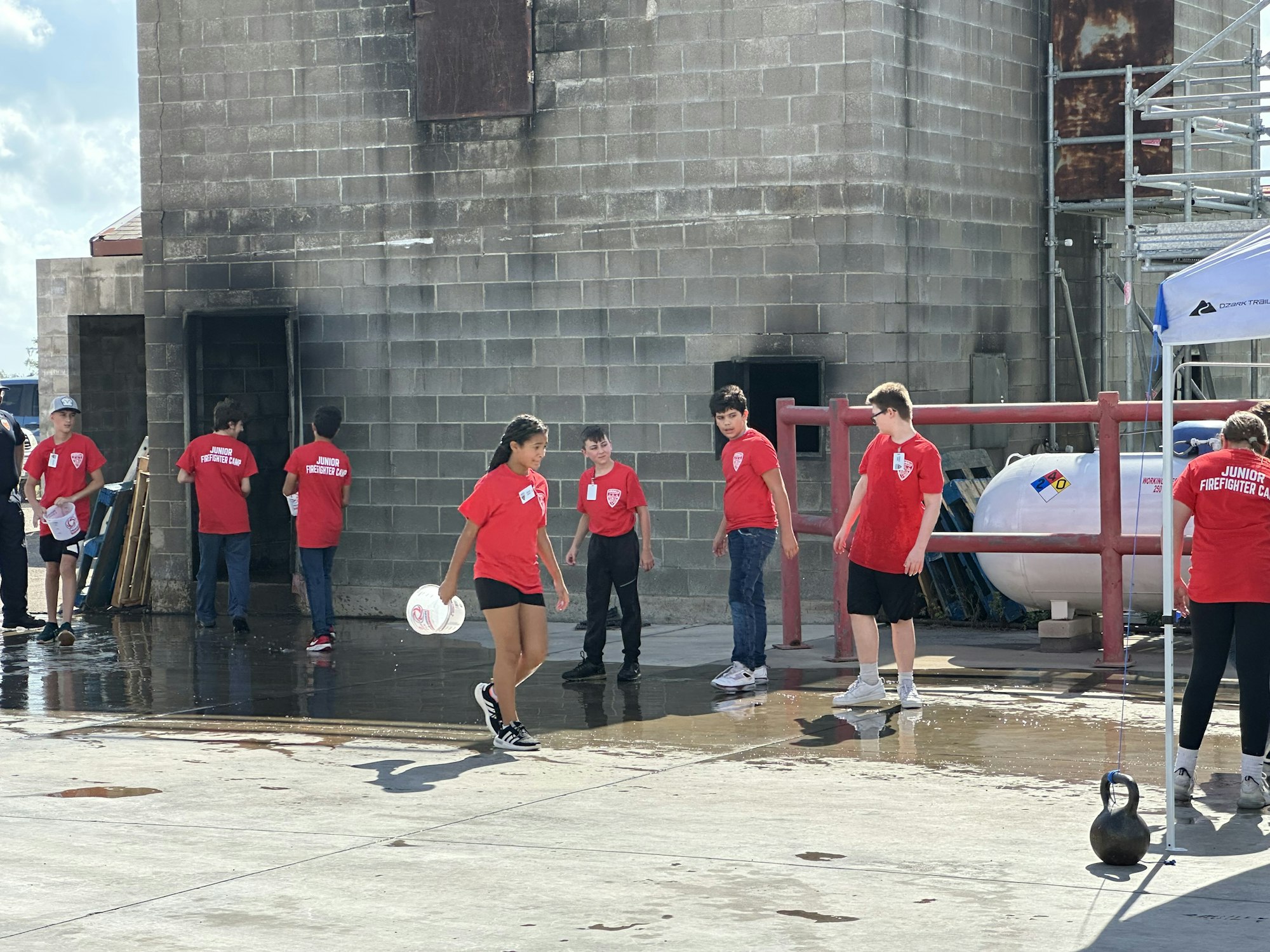 A group of young people wearing red "Junior Firefighter Camp" shirts are gathered near a concrete building and equipment.
