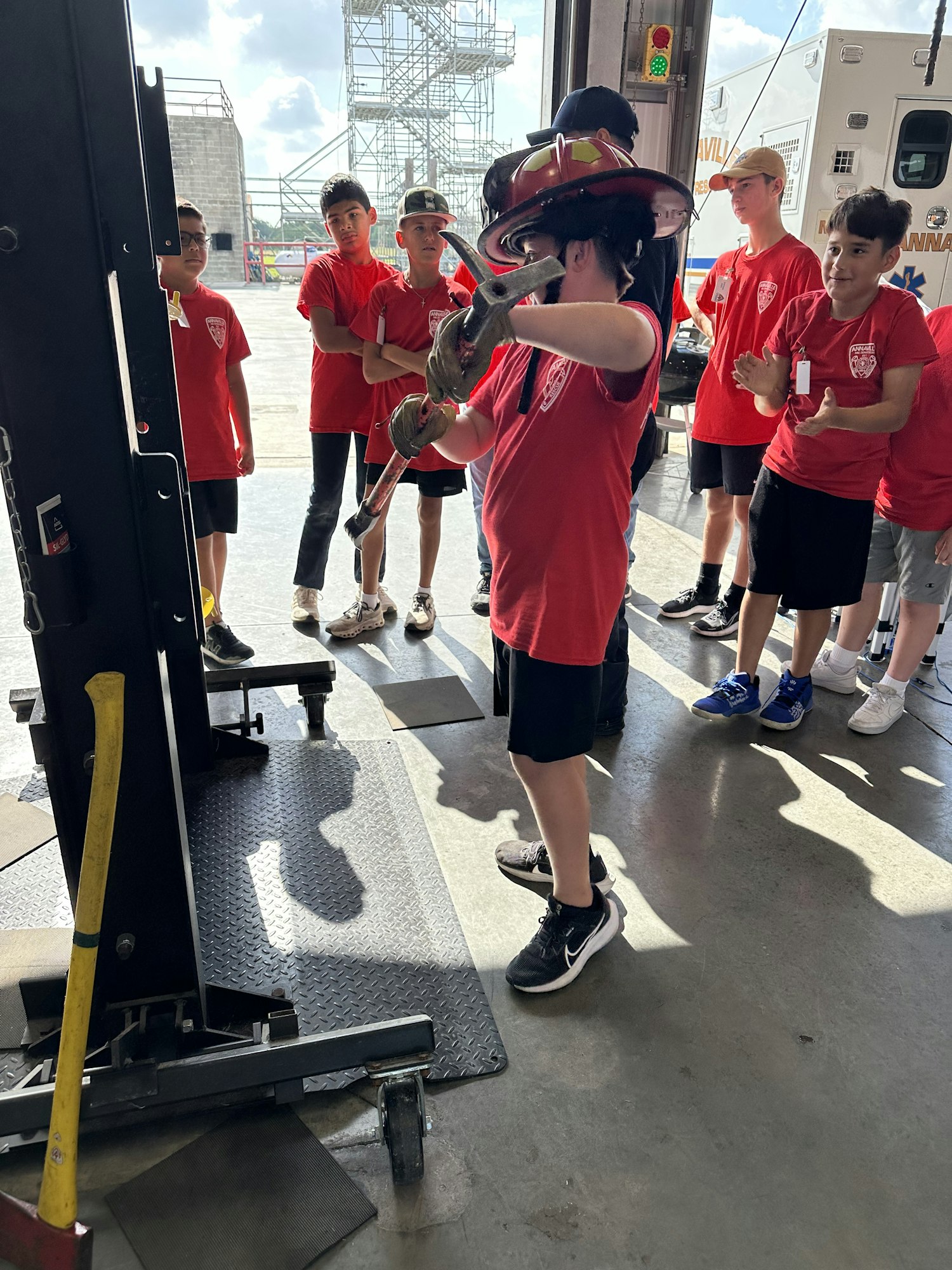 Group of people in red shirts, one wearing a firefighter helmet, operating equipment inside a garage-like space.
