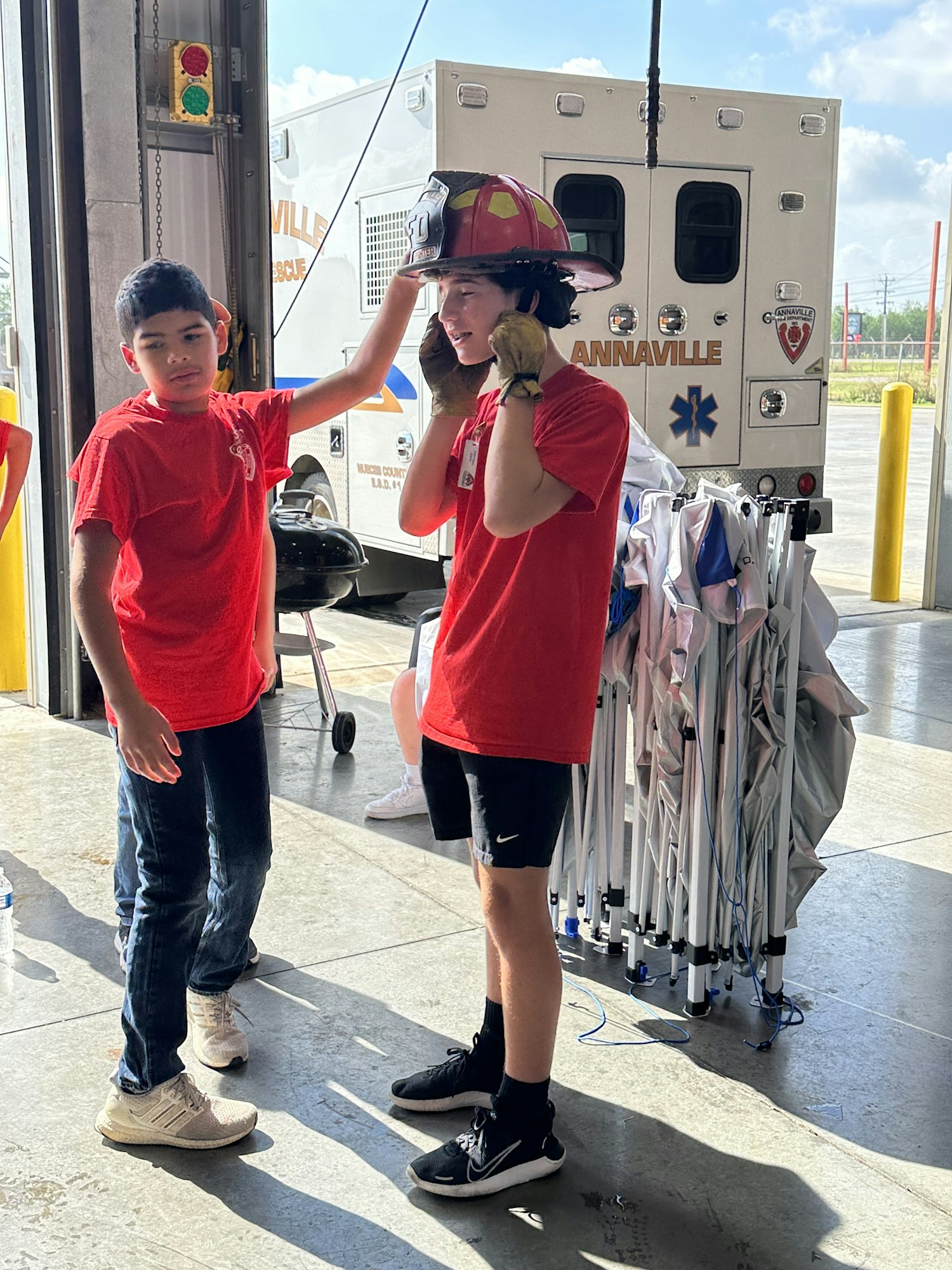 Two kids in red shirts; one wears a fire helmet. They're standing by a parked ambulance with folded canopies in the background.