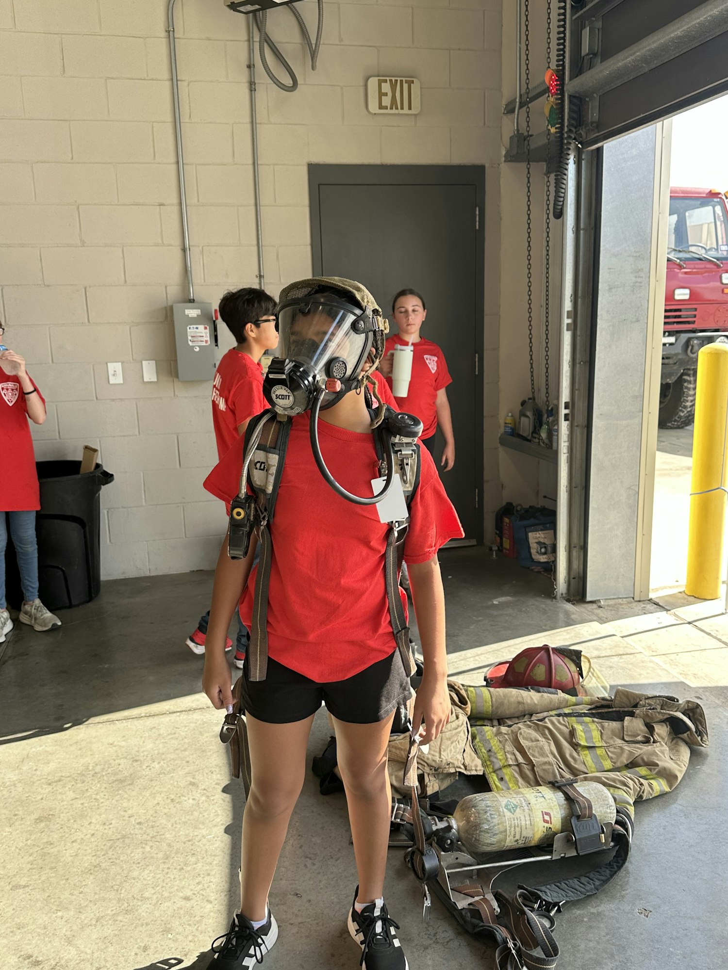 A person wearing firefighter gear indoors, with others in red shirts and a firefighting outfit on the ground.