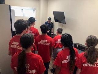 A group of kids in "Junior Firefighter Camp" shirts listen to an instructor in a small room with a TV.