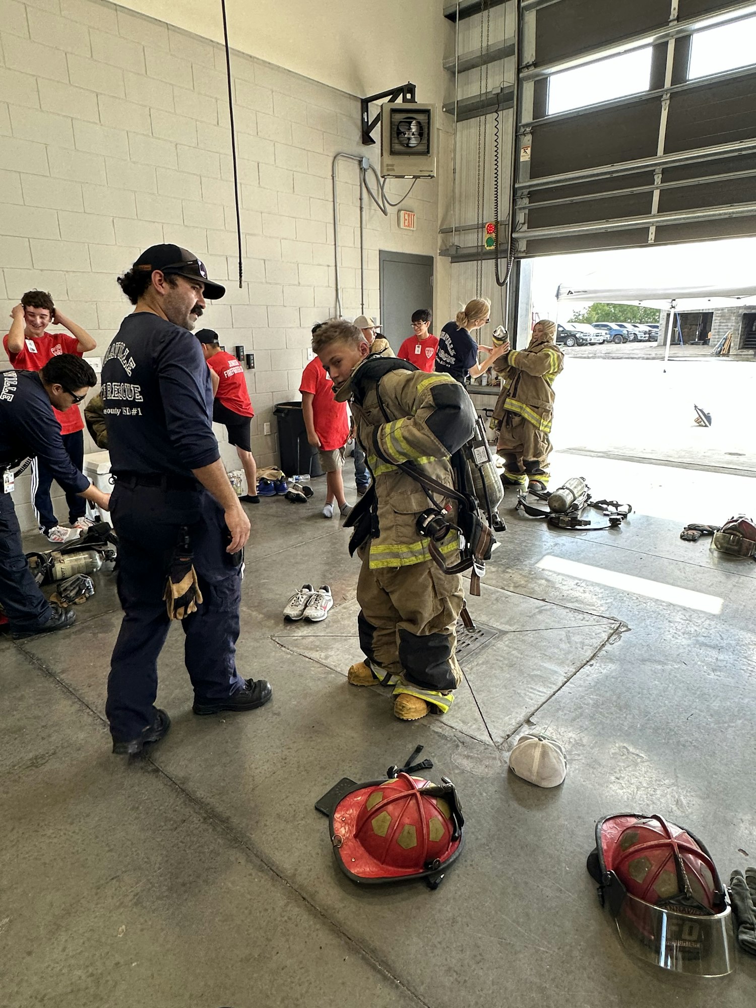 Firefighters stand in a station, with equipment on the ground and people in red shirts nearby.