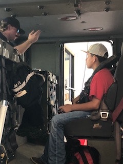 Two people sitting inside a vehicle, one in a red shirt and cap, the other in dark clothing gesturing upward.