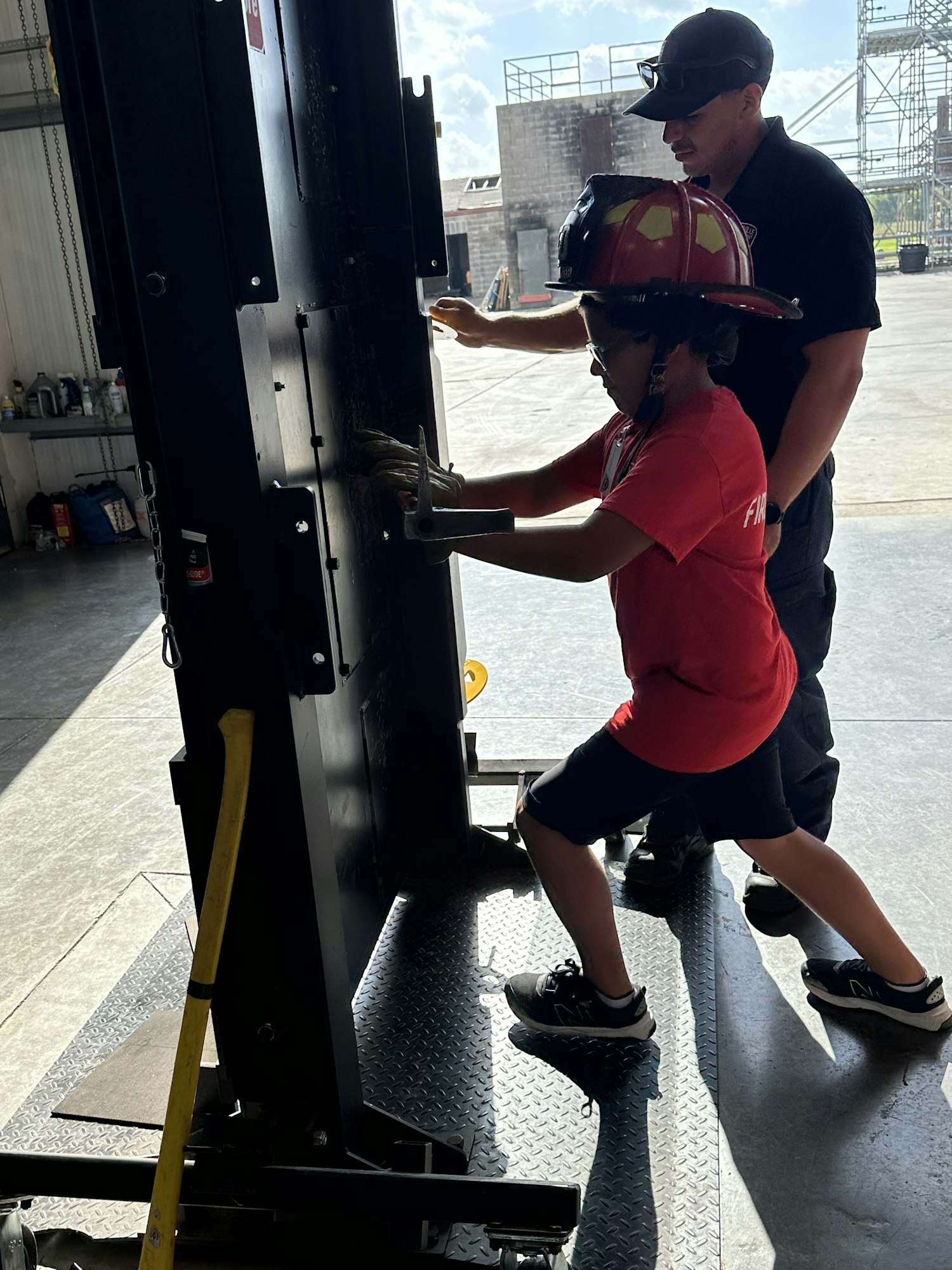A person in a fire helmet using a tool on a large metal structure, supervised by another person.