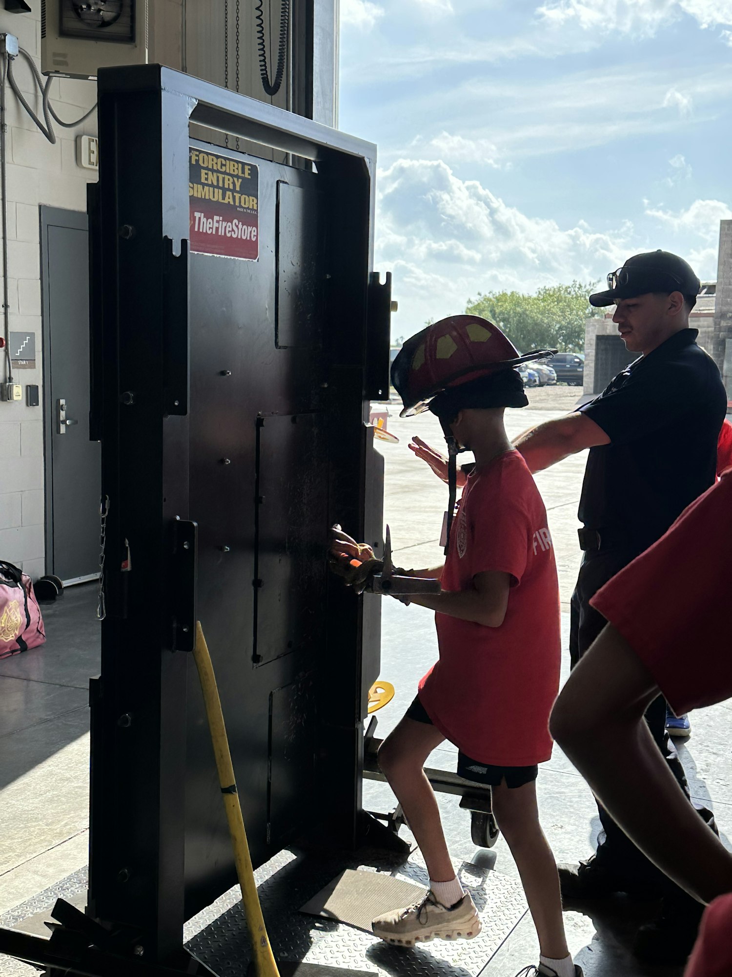 A person is using a forcible entry simulator under guidance, wearing a firefighter helmet and red shirt.