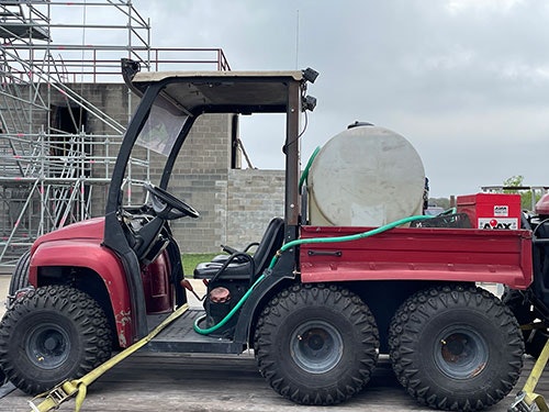 A red utility vehicle with six wheels, carrying a large tank, parked in front of scaffolding and a partially constructed building.