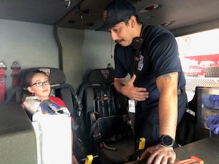 A firefighter talks to a child inside a fire truck, explaining something with a hand gesture.