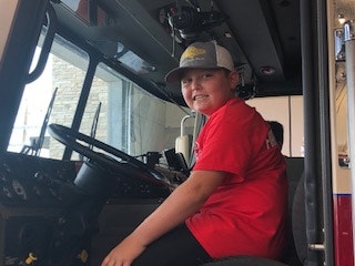 A child in a red shirt and cap sits in the driver's seat of a vehicle, possibly a truck or fire engine.