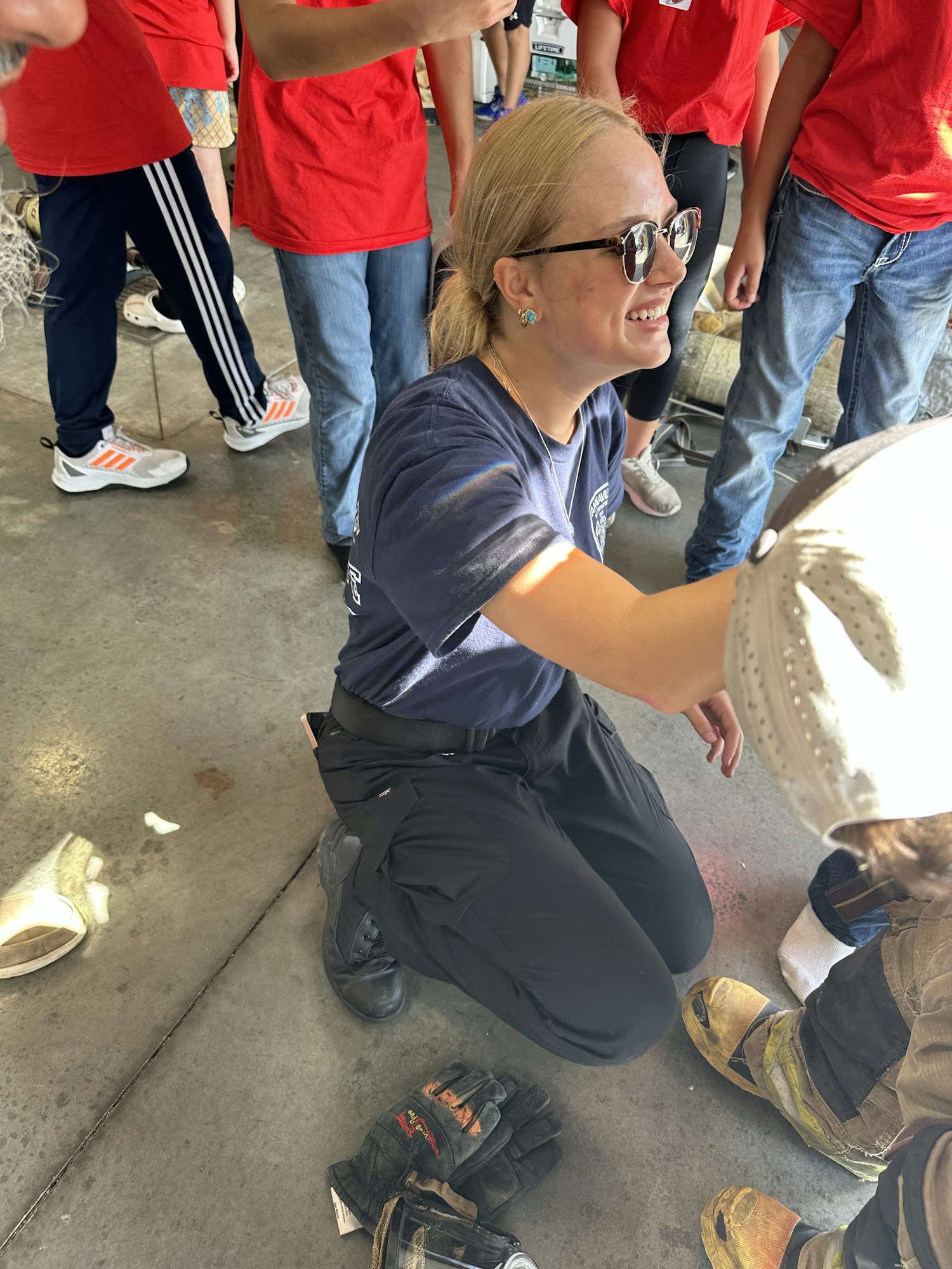 A person is kneeling and interacting with a group, including a firefighter with gear and gloves on the floor nearby.