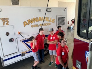 A group of people in red shirts stands near an "ANNAVILLE FIRE-RESCUE" vehicle inside a garage.
