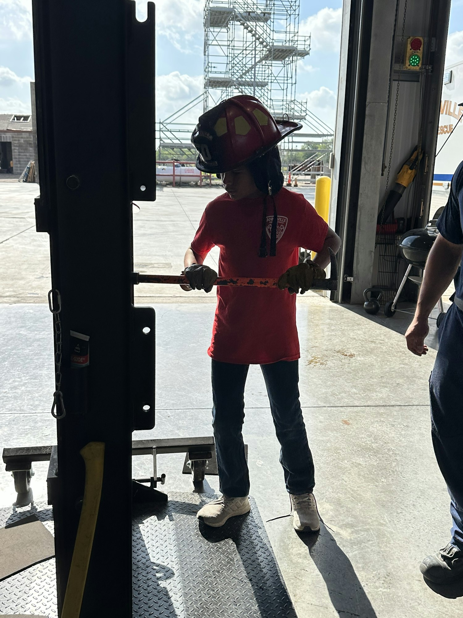A firefighter in a red helmet and shirt is using a tool inside a garage with industrial structures in the background.