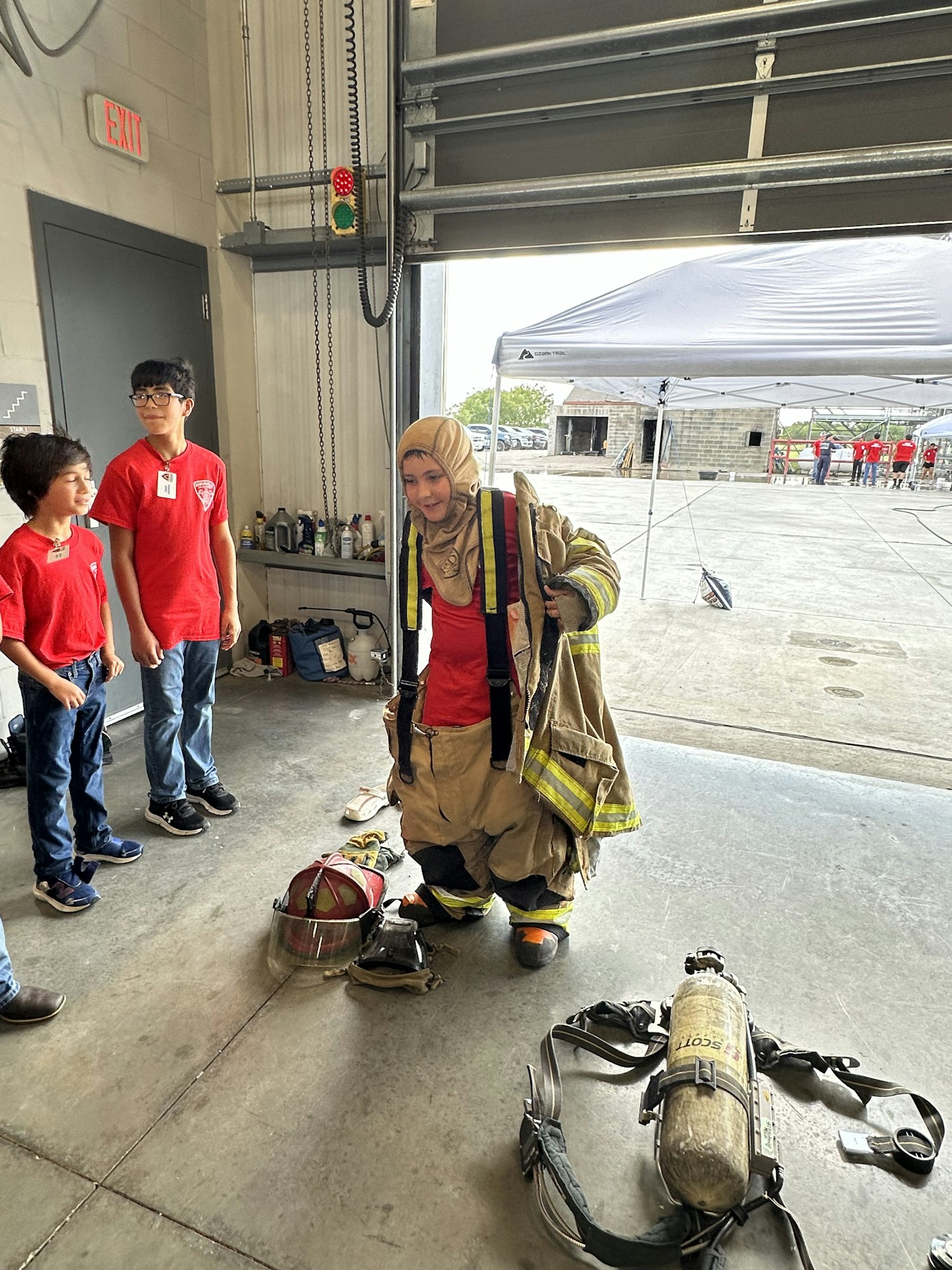 A woman in firefighting gear kneels indoors beside two kids in red shirts; firefighting equipment is on the floor nearby.