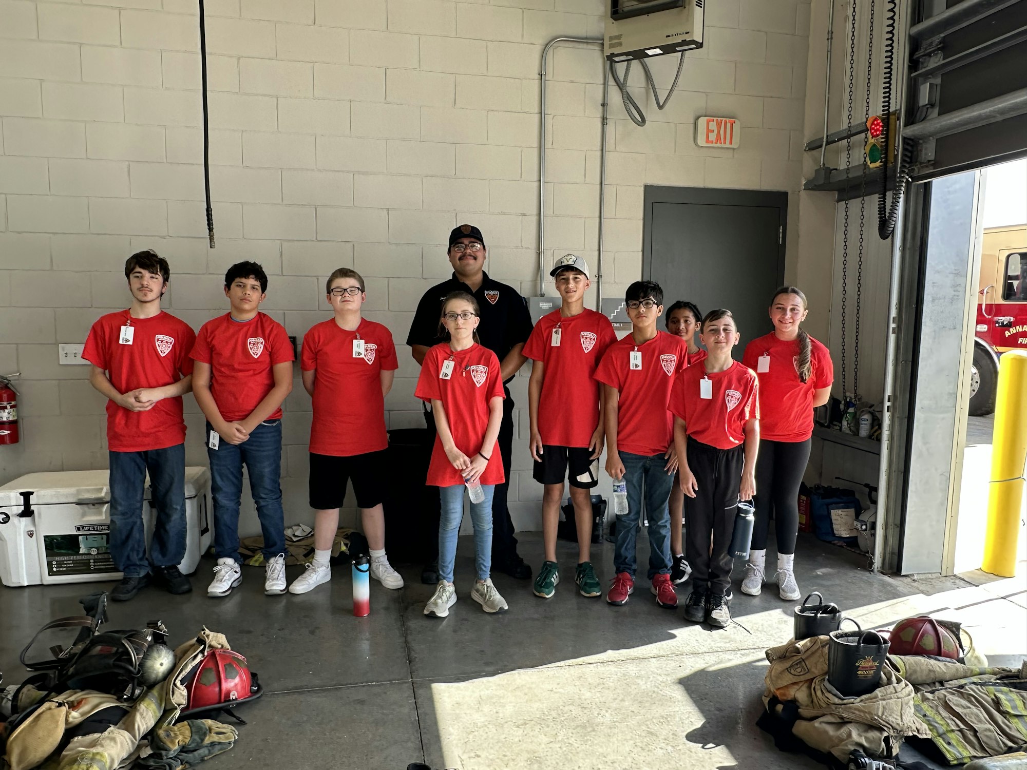 A group of kids in matching red shirts pose inside a garage with fire equipment on the floor.