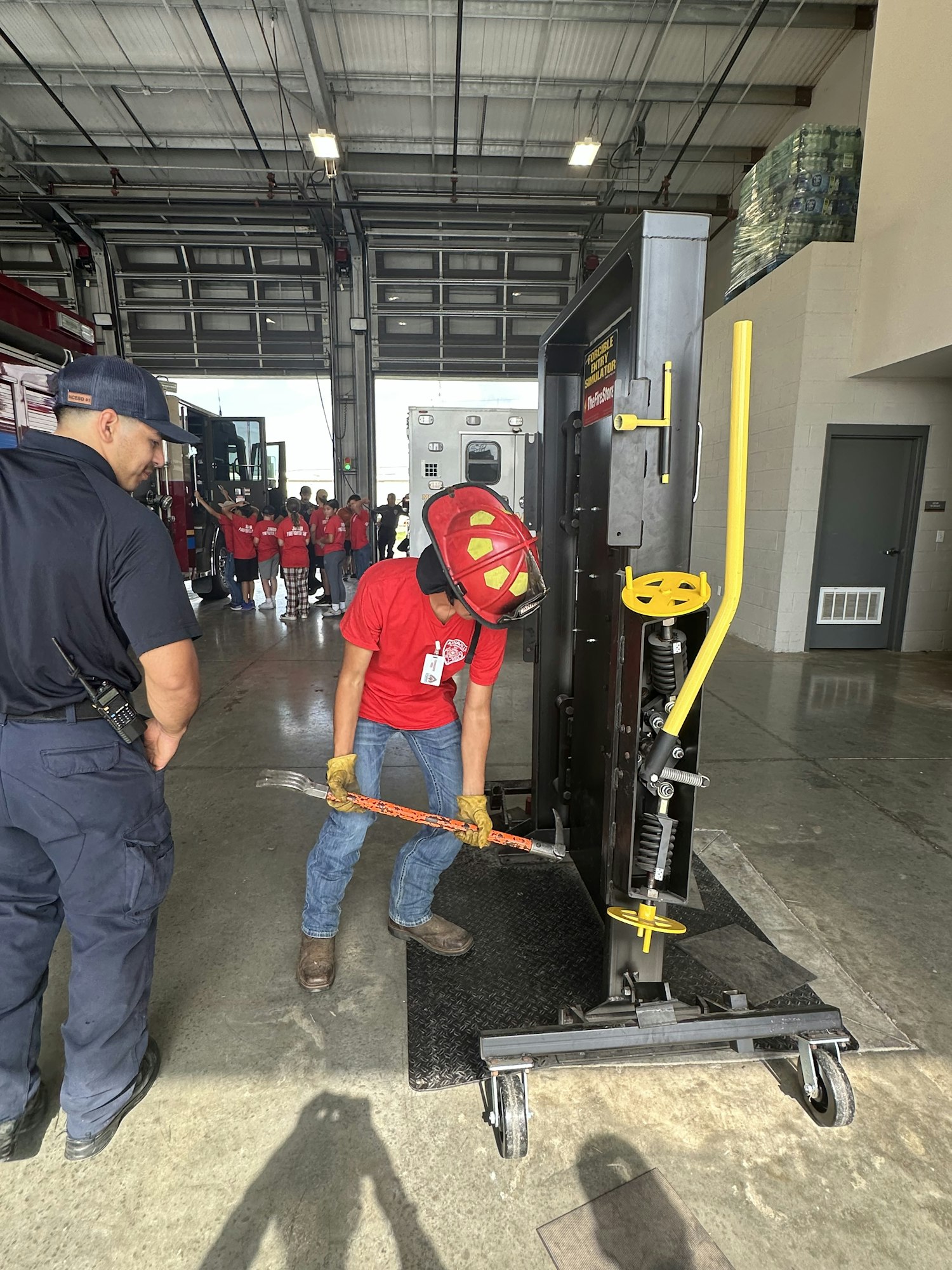 A person in a red helmet and shirt uses a tool on equipment inside a large garage, observed by another person in a blue cap.