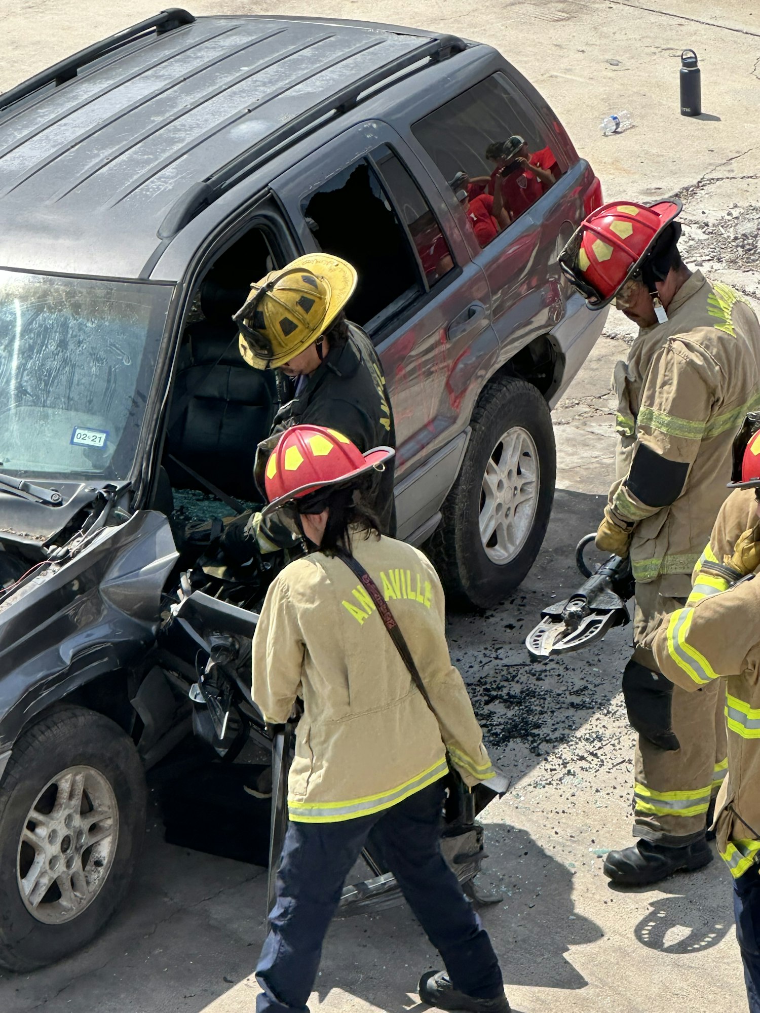 Firefighters in gear at a car accident scene, examining a damaged vehicle with broken windows.