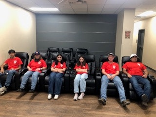 Six people in red shirts and jeans are sitting on black recliner chairs in a room with beige walls and wood floors.