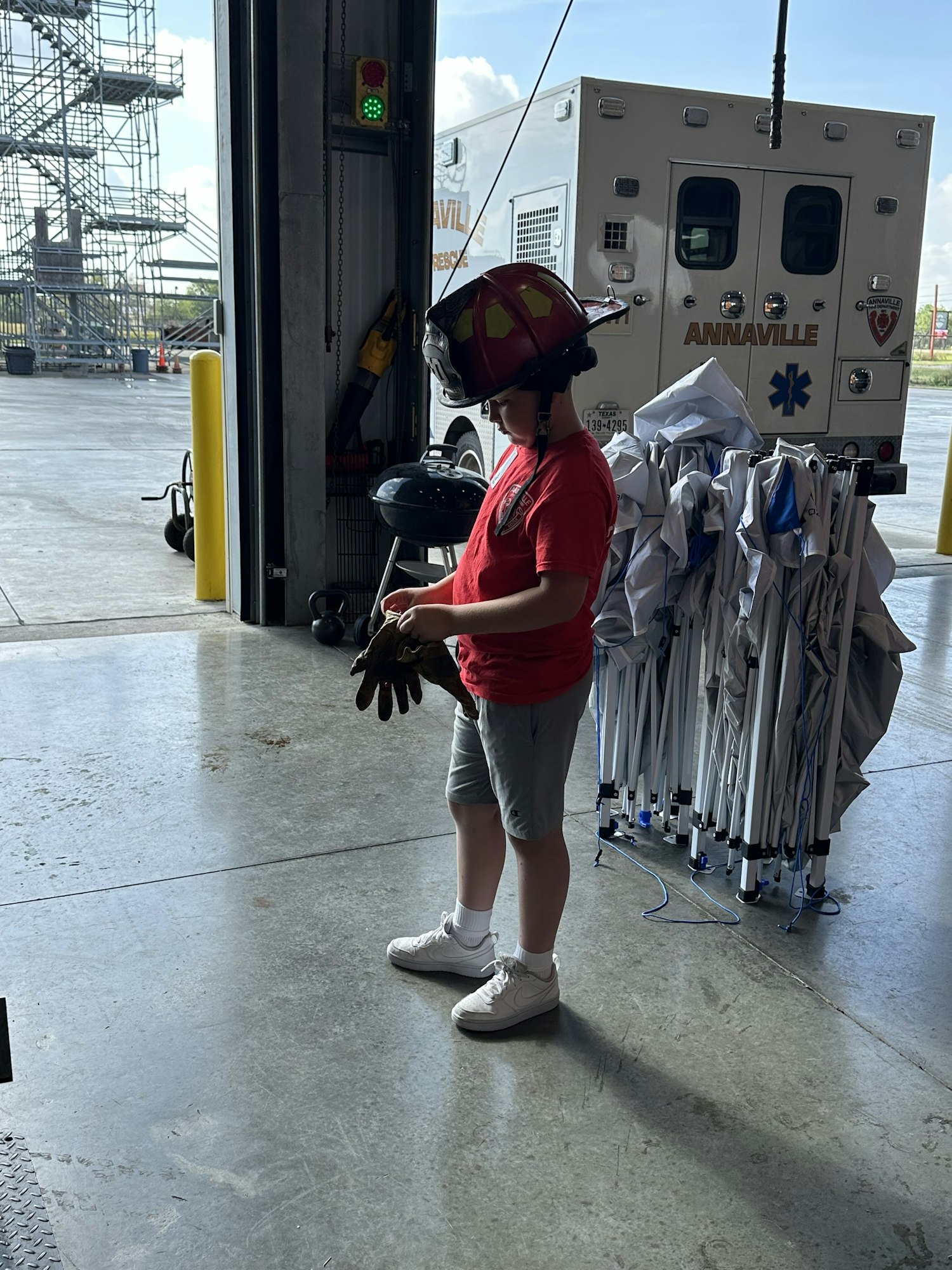 A person in a red helmet and gloves stands near an ambulance and equipment inside a building.
