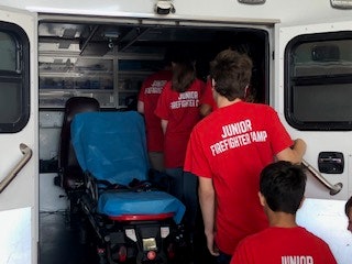 People wearing "JUNIOR FIREFIGHTER CAMP" shirts entering an ambulance with a stretcher inside.