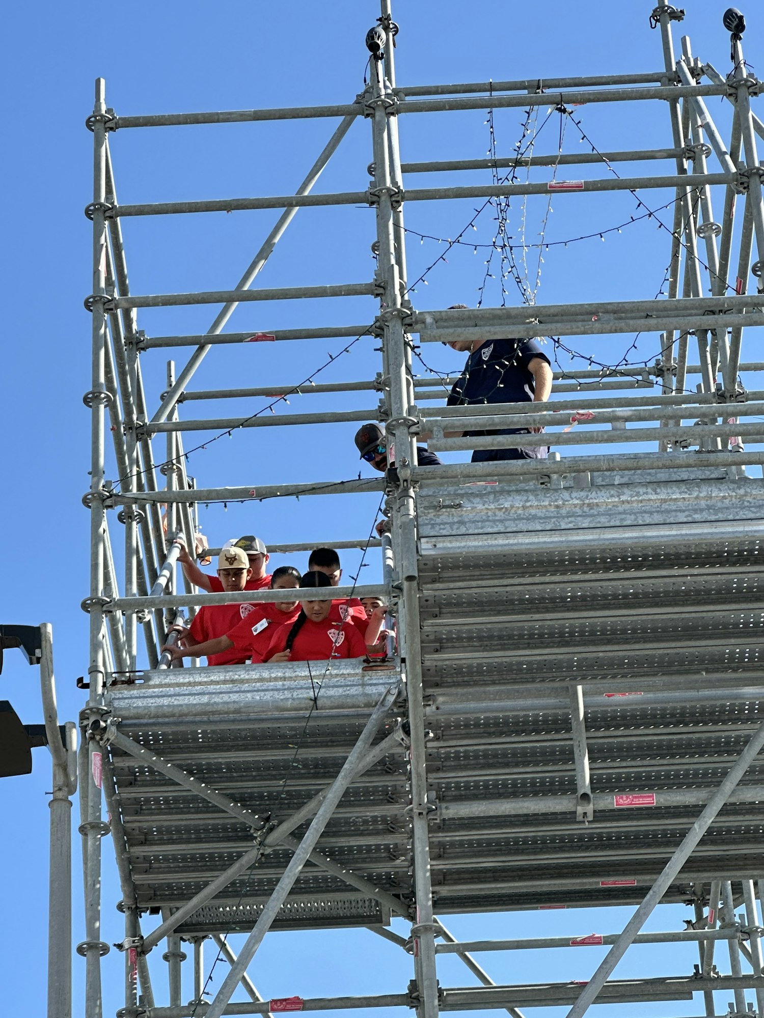 People in red shirts climb metal scaffolding under a clear blue sky.