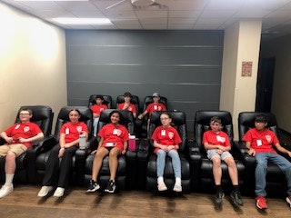 A group of nine people sitting in reclining chairs, all wearing red shirts with nametags, in a room with a gray wall.