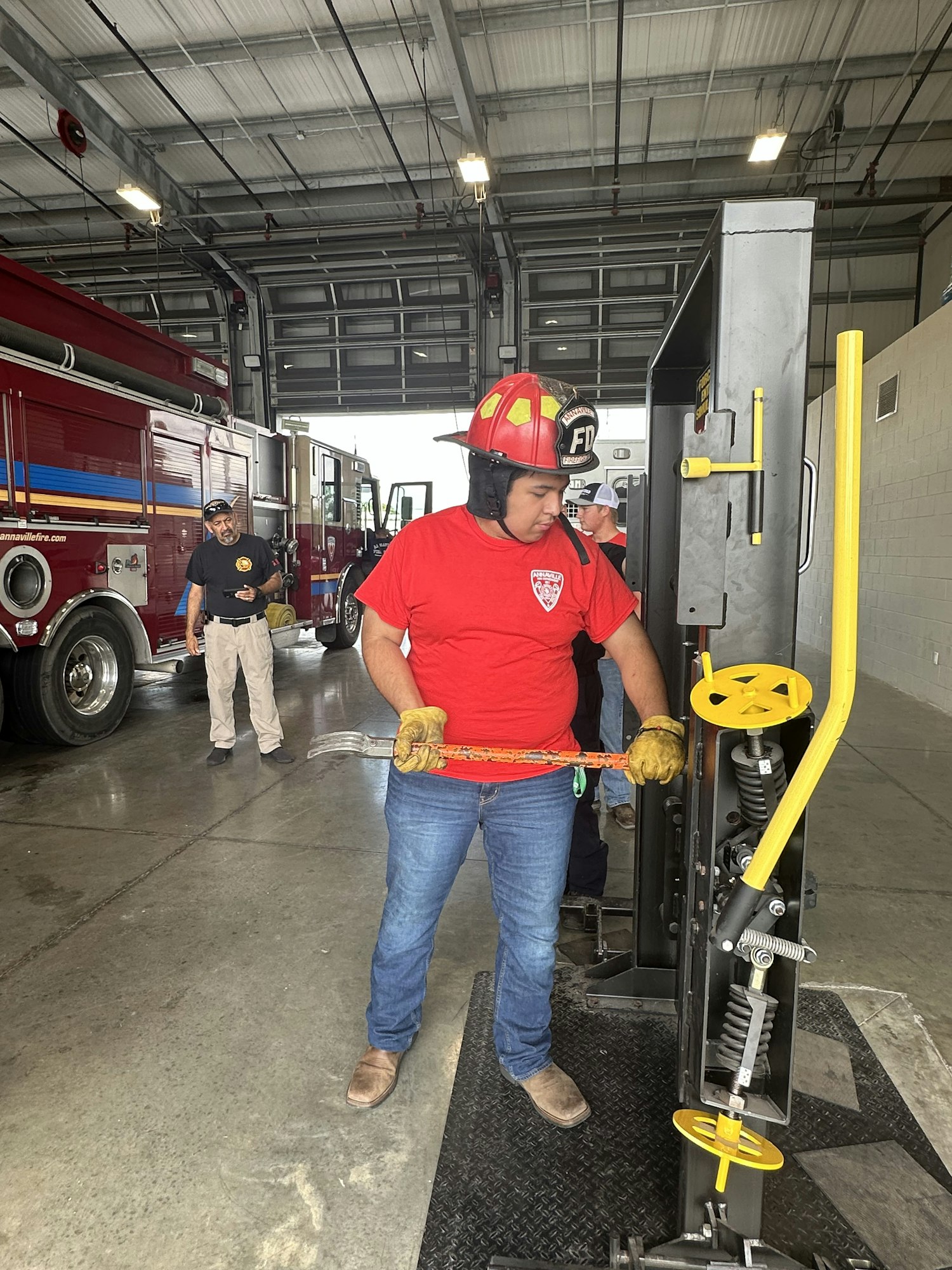 A person in a red helmet using a tool on a device, wearing gloves and jeans inside a garage with a fire truck visible.