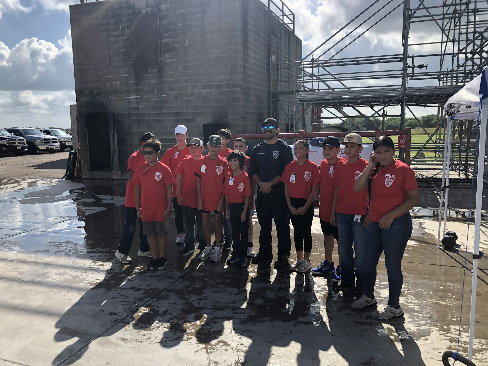 A group of people in matching red shirts stands outdoors near a building and scaffolding, with some cars visible in the background.