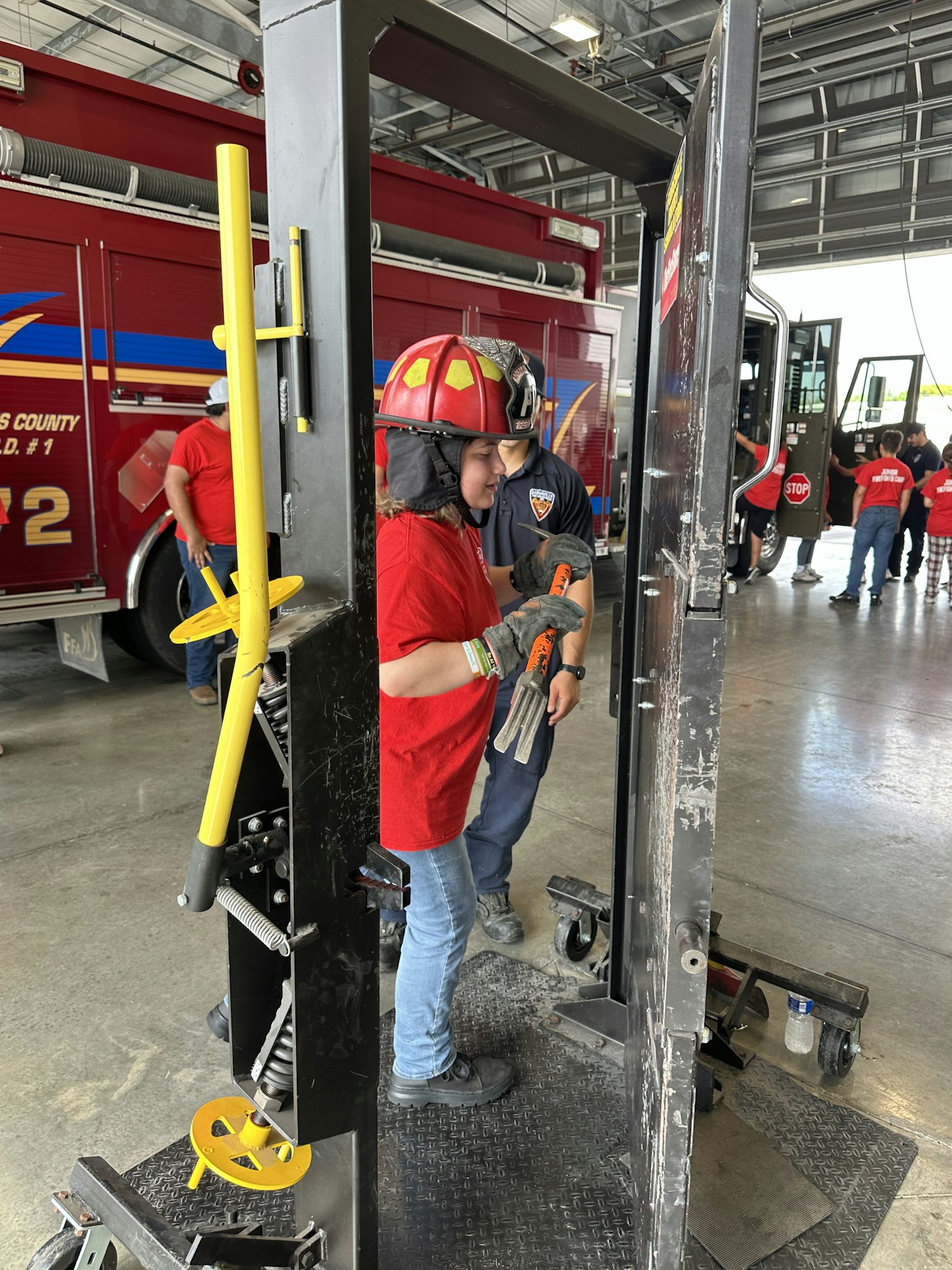 Two people in a fire station, one wearing a red firefighter helmet, using equipment, with fire trucks visible in the background.