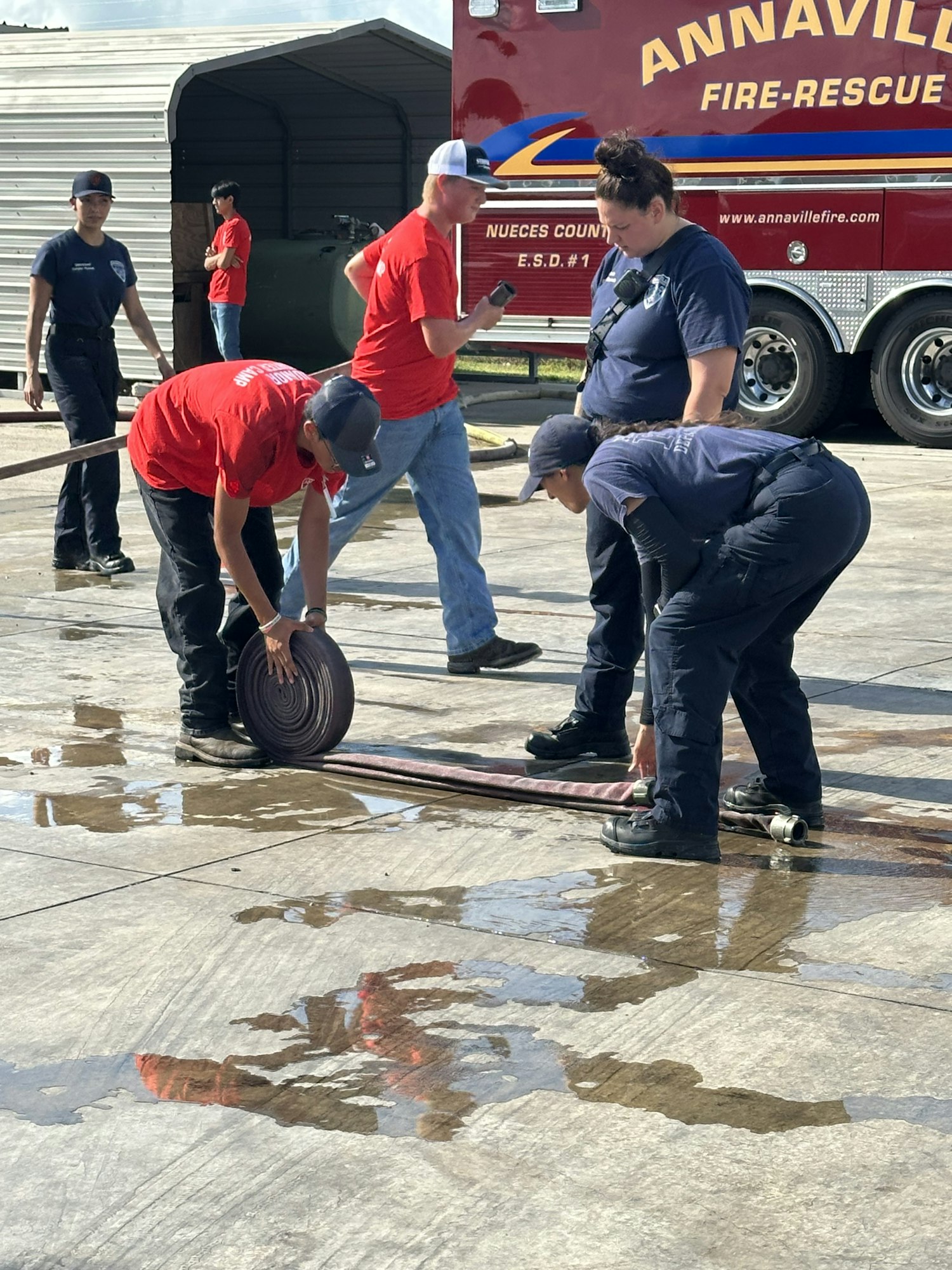Firefighters and trainees handling and rolling up a fire hose near a fire truck on a wet concrete surface.