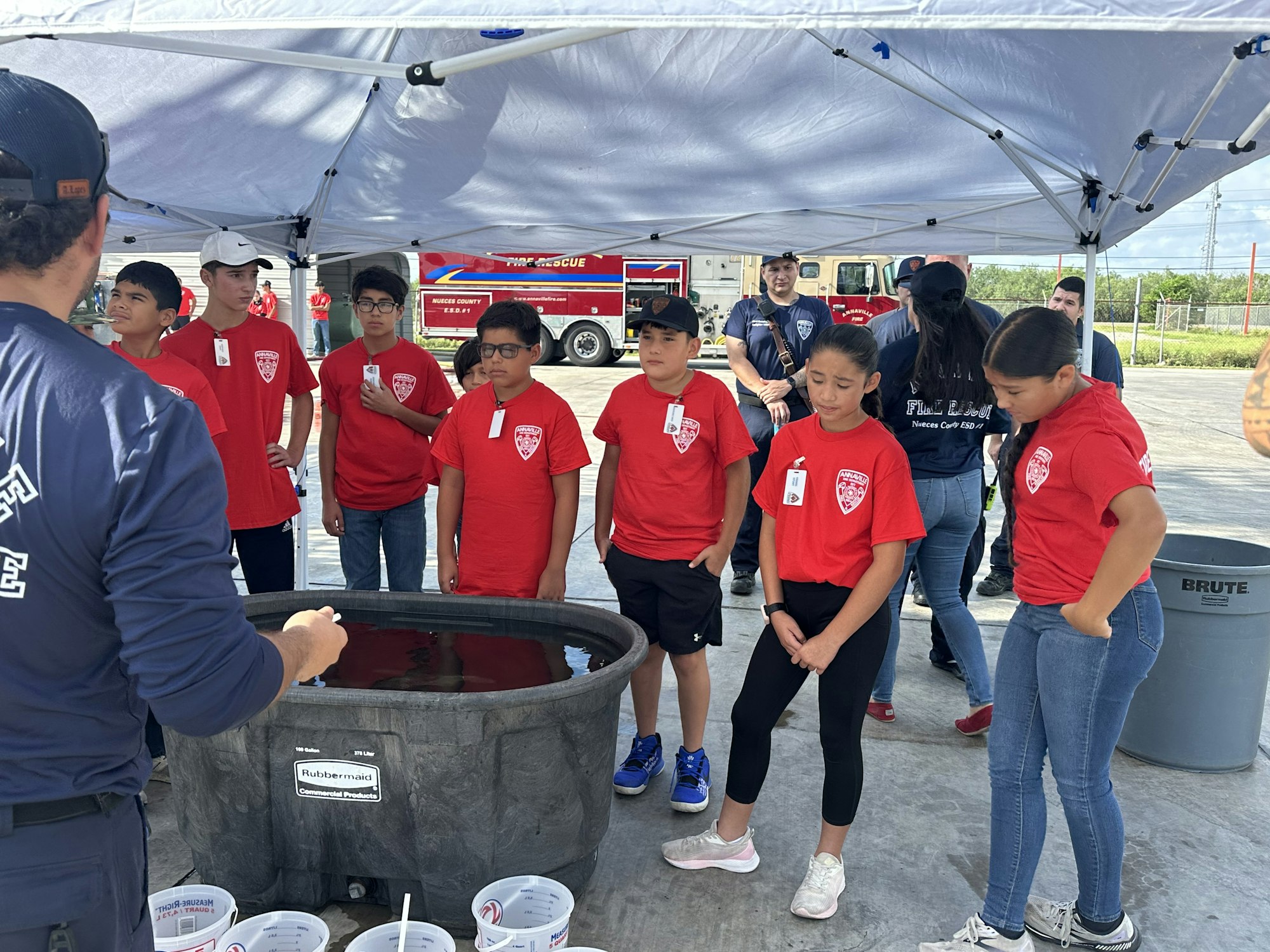 A group of kids in red shirts stand under a canopy, listening to a person beside a large container, with fire rescue visible in the background.