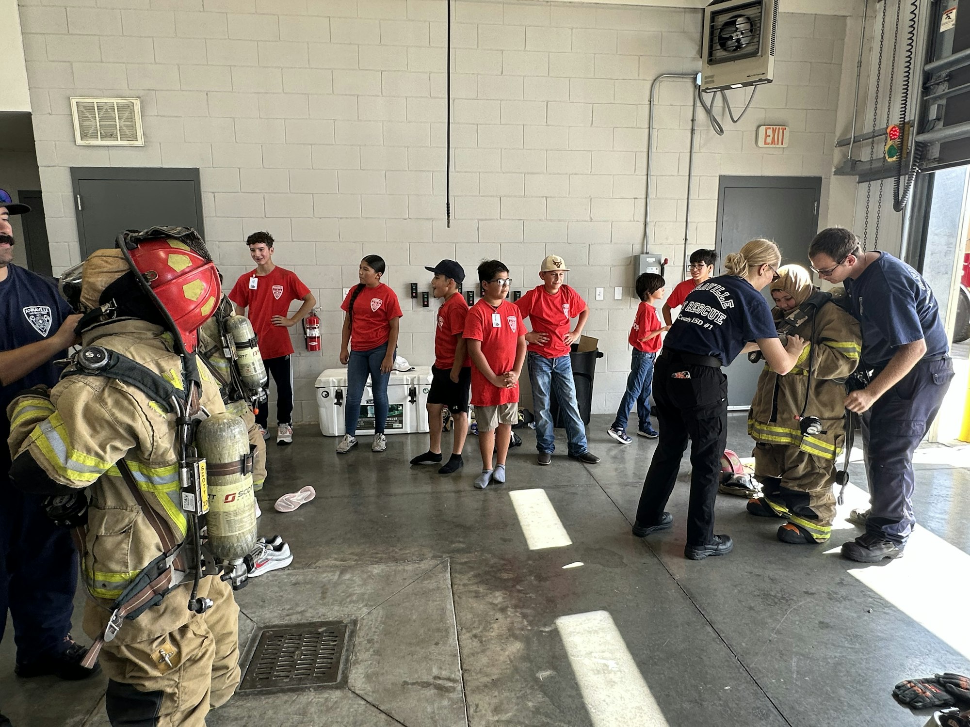 Firefighters helping kids try on gear during a station visit; children in red shirts watch and participate.