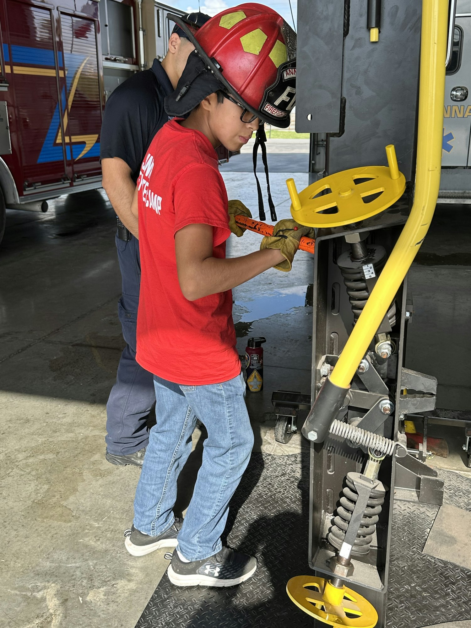 A person in a red shirt and helmet is using a tool, assisted by another person in a helmet, near firefighting equipment.