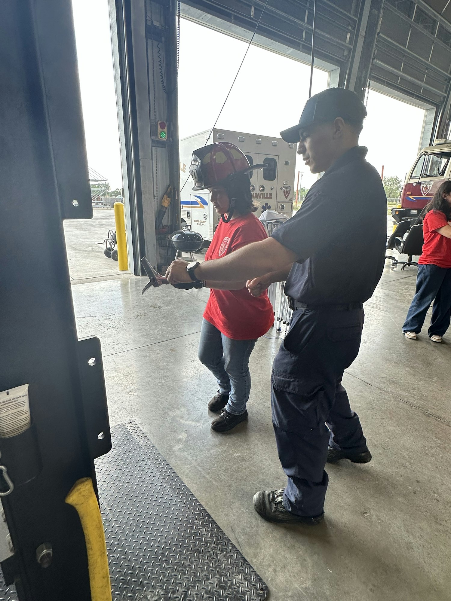 Two people standing in a garage; one in red wearing a firefighter helmet, the other in dark uniform; an ambulance is visible outside.