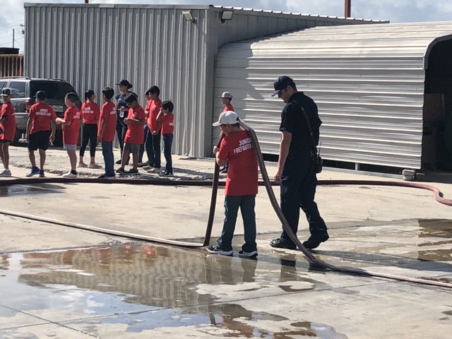 Group of kids in red shirts lined up, participating in a firefighting activity with a firefighter, using a hose.